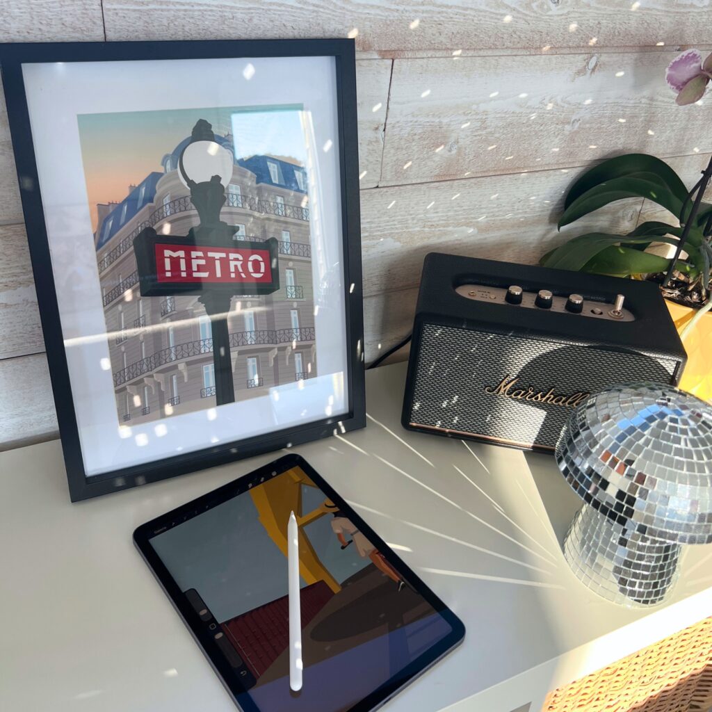 Affiche du Métro parisien encadré, posé sur un meuble blanc sur lequel se trouve un IPad ouvert sur une illustration de Nekae Studio