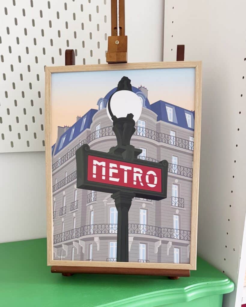 Affiche encadrée représentant le panneau du métro parisien, posée sur un chevalet d'artiste en bois placé sur une commode.